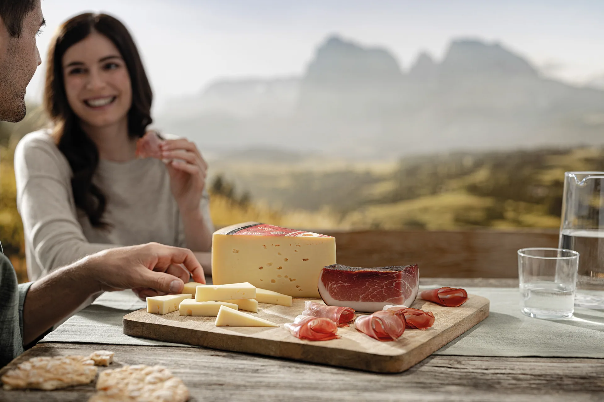 Paar genie&szlig;t K&auml;se und Speck auf einem Holzbrett vor Bergkulisse